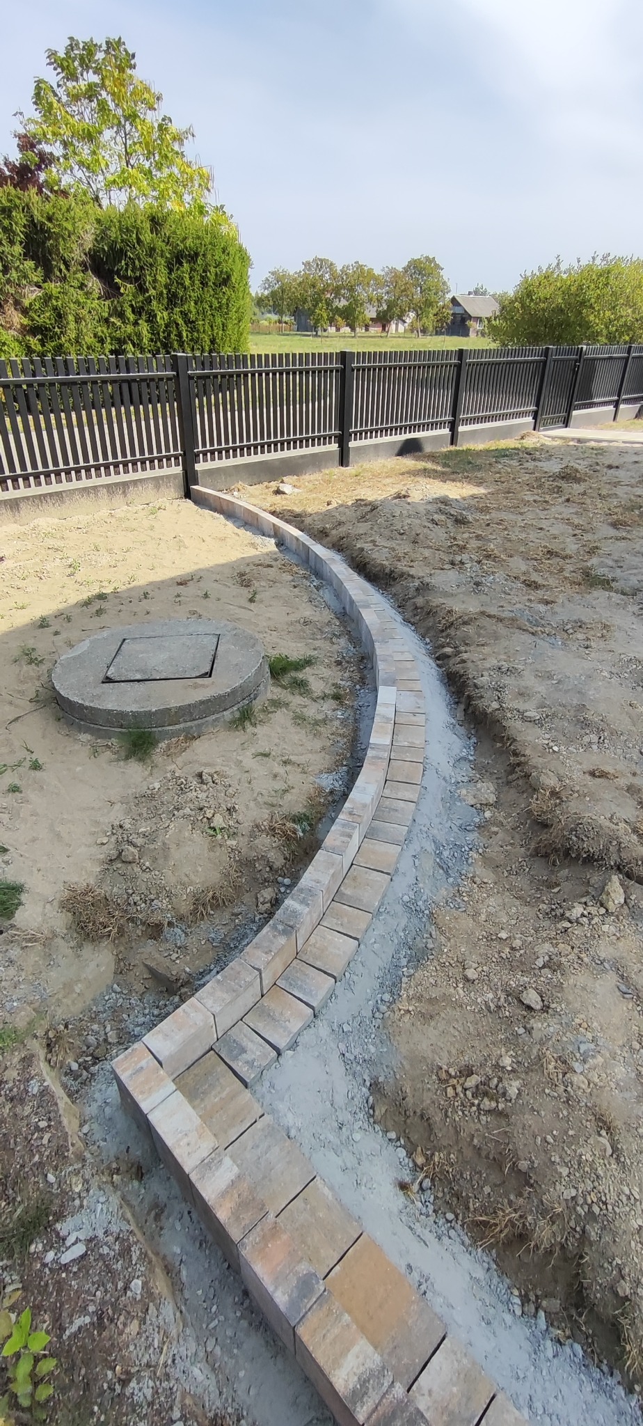 Krawężnik z kostki brukowej ułożony łukiem w Tarnogrodzie. Obok studzienka kanalizacyjna, w tle czarny płot i zieleń.