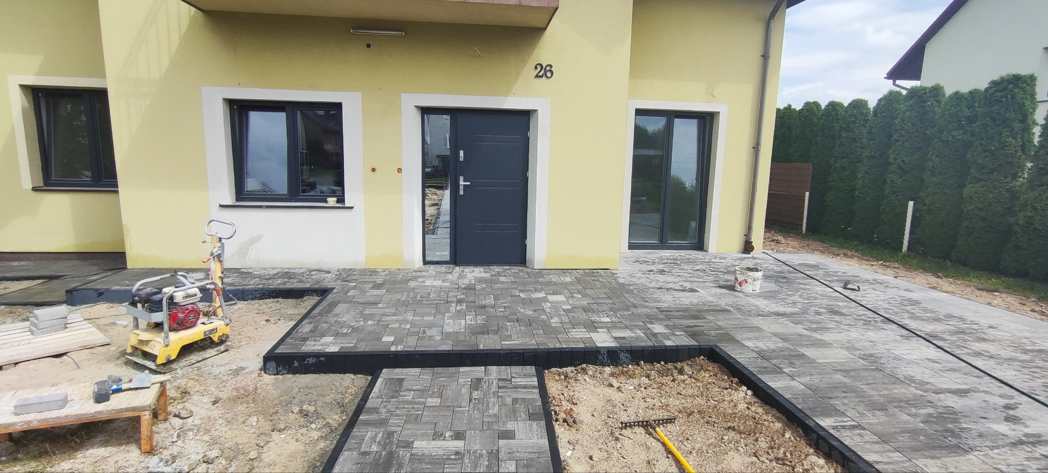 Fragment posesji z częściowo ułożoną kostką brukową w odcieniach szarości. Widoczny dom z numerem 26, narzędzia brukarskie i maszyna do ubijania.