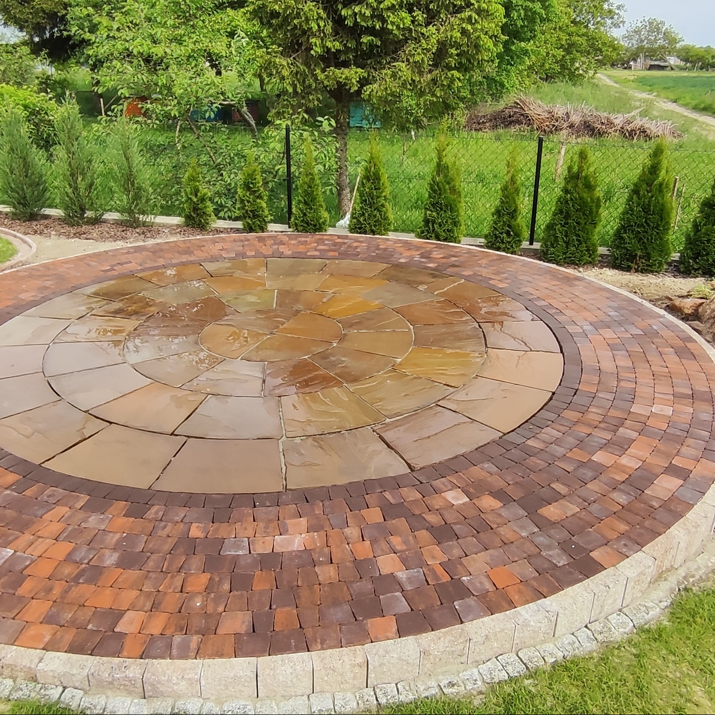 Okrągła, wielokolorowa nawierzchnia z kostki brukowej w ogrodzie, z obrzeżem z drobnej kostki i zielenią w tle. Układ radialny z kamieniami o różnych odcieniach brązu.