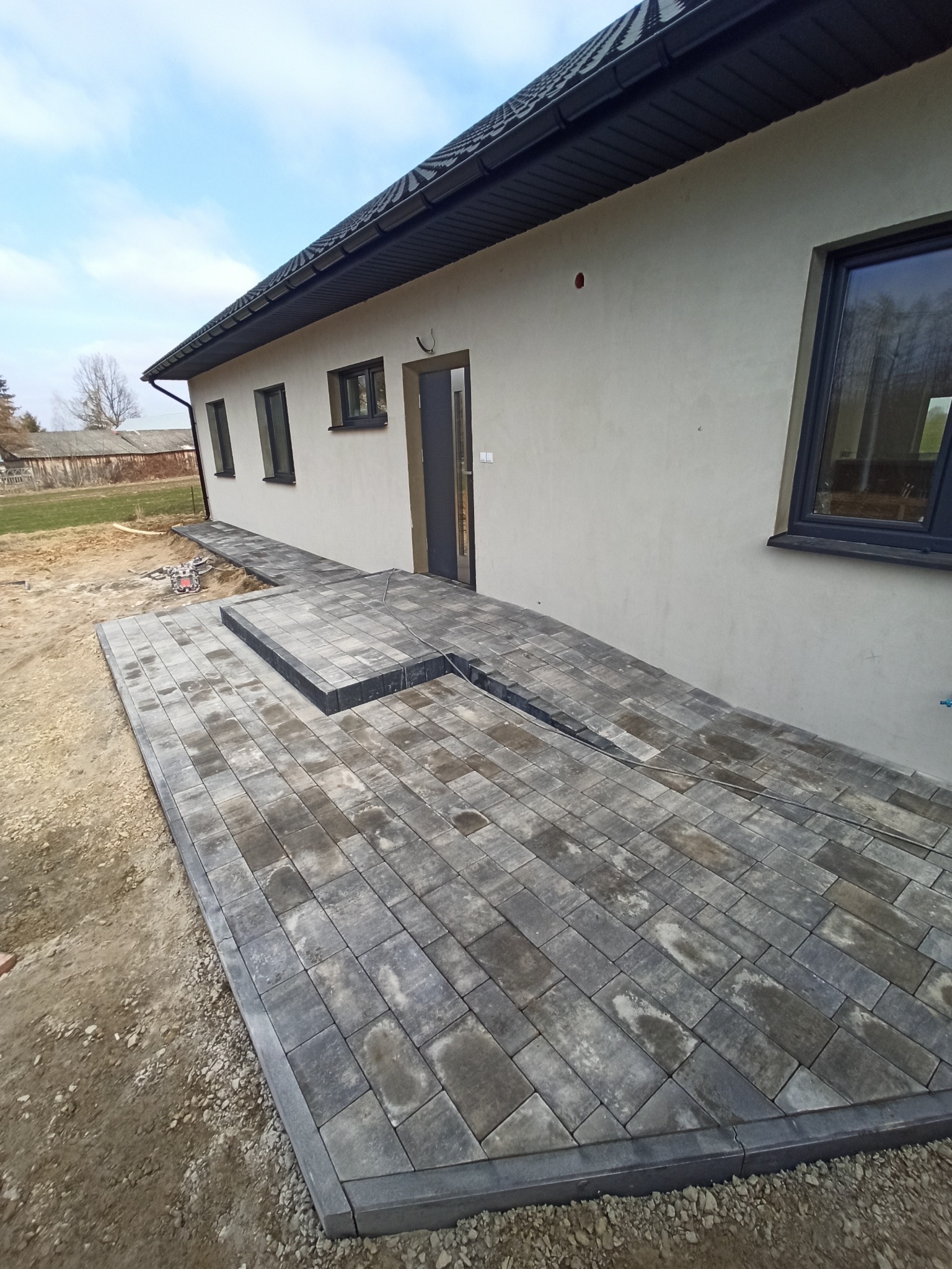 Ułożona szara kostka brukowa tworząca taras i ścieżkę przy jasnym domu z ciemnymi oknami i drzwiami, widoczne niedokończone obrzeża i przygotowany teren.