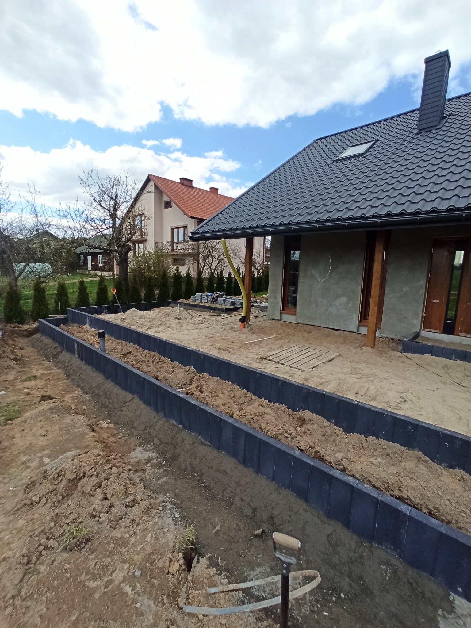 Przygotowanie terenu pod trawnik lub ogród: wytyczone krawężniki z wypełnieniem ziemią, widoczny fragment domu z ocieploną elewacją, narzędzia ogrodnicze na pierwszym planie.