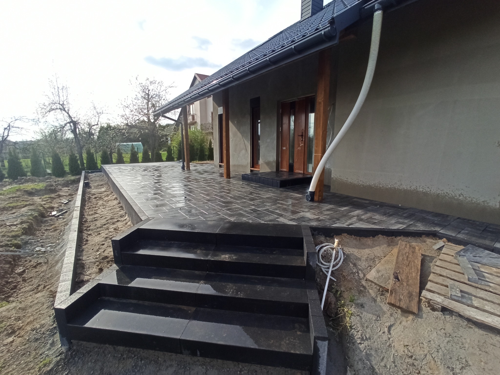 Nowo ułożona, mokra od deszczu, czarna kostka brukowa tworząca schody i taras przed domem w Tarnogrodzie. Widoczne drewniane podpory zadaszenia i fragment ogrodu w tle.