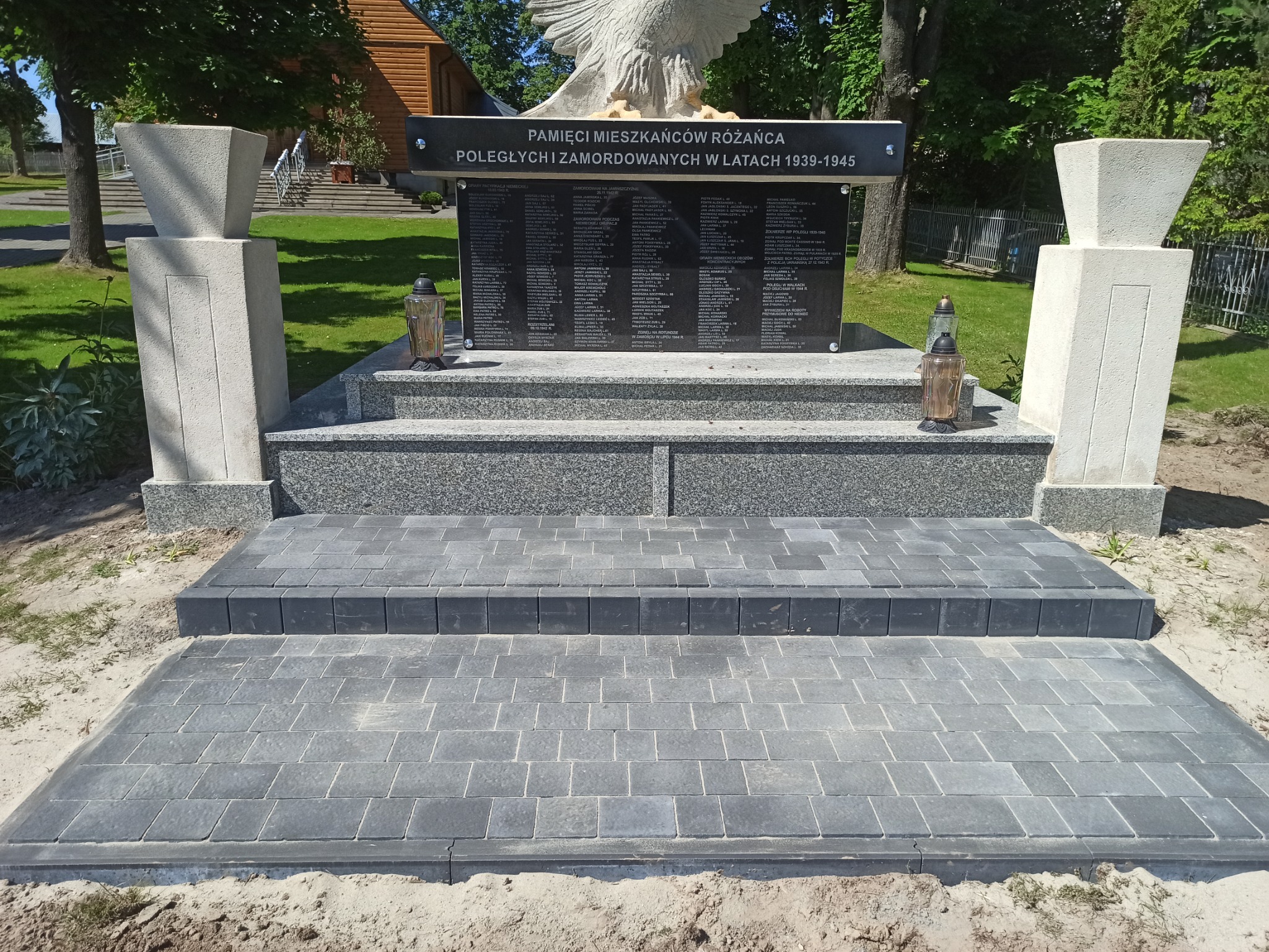 Nowa, szara kostka brukowa ułożona przed pomnikiem upamiętniającym ofiary wojny, z widocznymi dwoma lampionami i elementami architektonicznymi w tle.