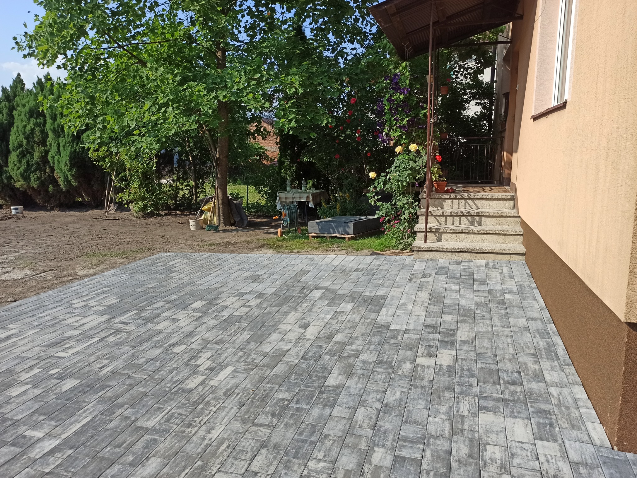 Nowo ułożona, szara kostka brukowa w jodełkę na podwórku przed domem z beżową elewacją, widoczne schody wejściowe i fragment ogrodu z zielenią.