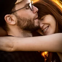 Młoda para w bliskim uścisku na tle zachodzącego słońca; mężczyzna w okularach i czapce z daszkiem z korkowym elementem, kobieta z uśmiechem i aparatem ortodontycznym.