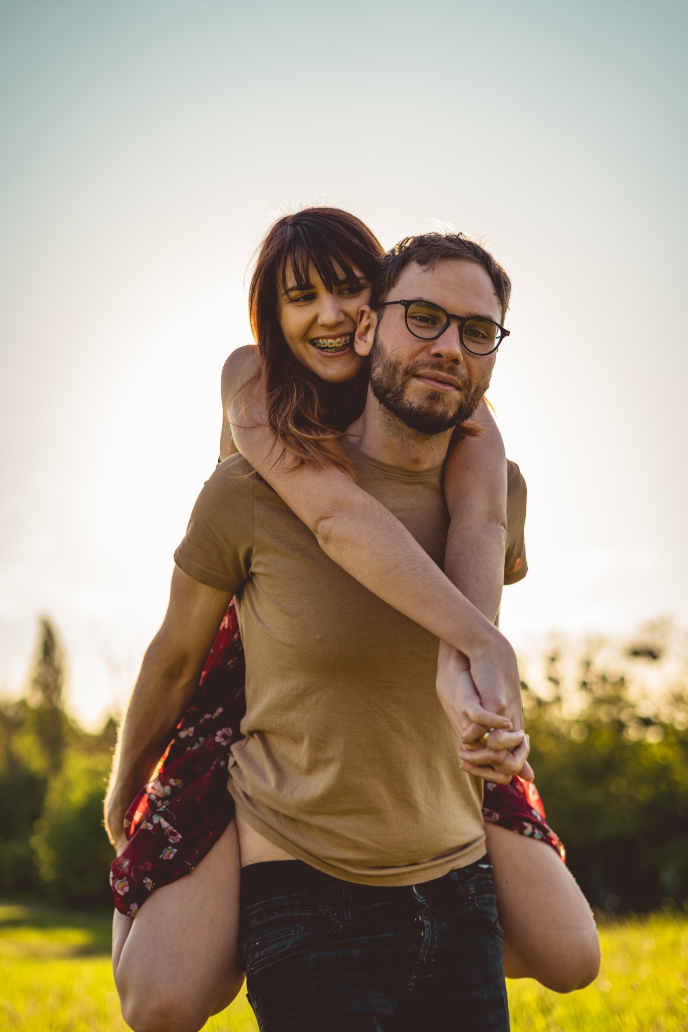 Uśmiechnięta kobieta z aparatem ortodontycznym na barana u mężczyzny w okularach, na tle rozmytej zieleni i zachodzącego słońca.