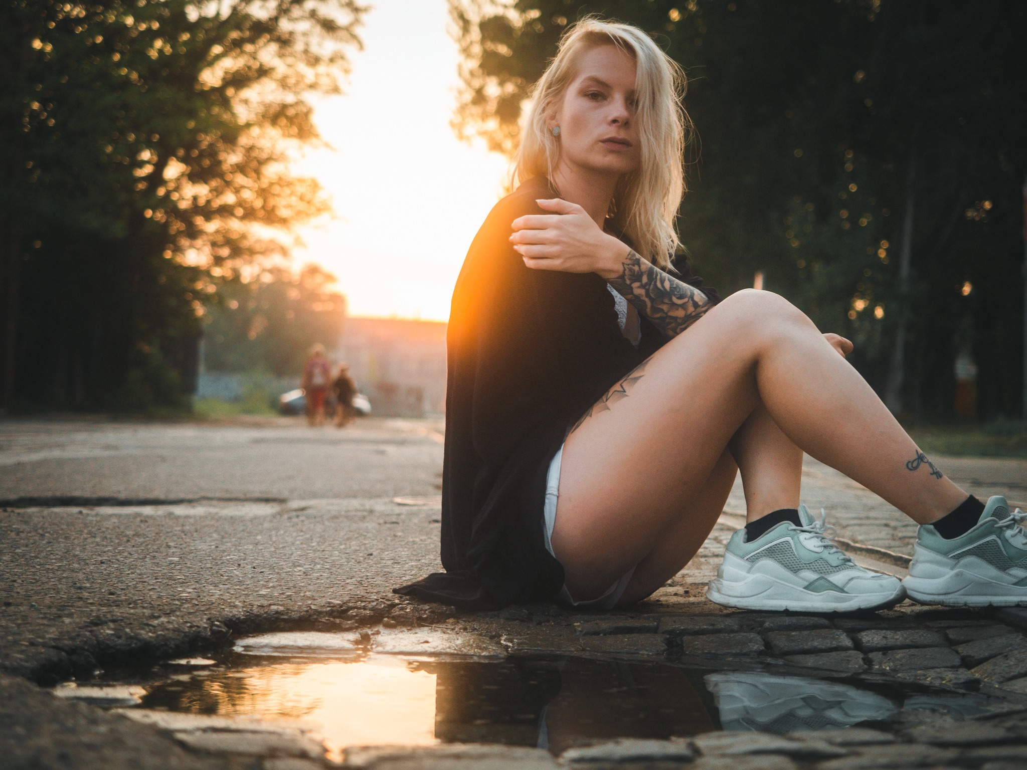 Kobieta z tatuażami siedzi na asfalcie obok kałuży, w tle drzewa i zachodzące słońce