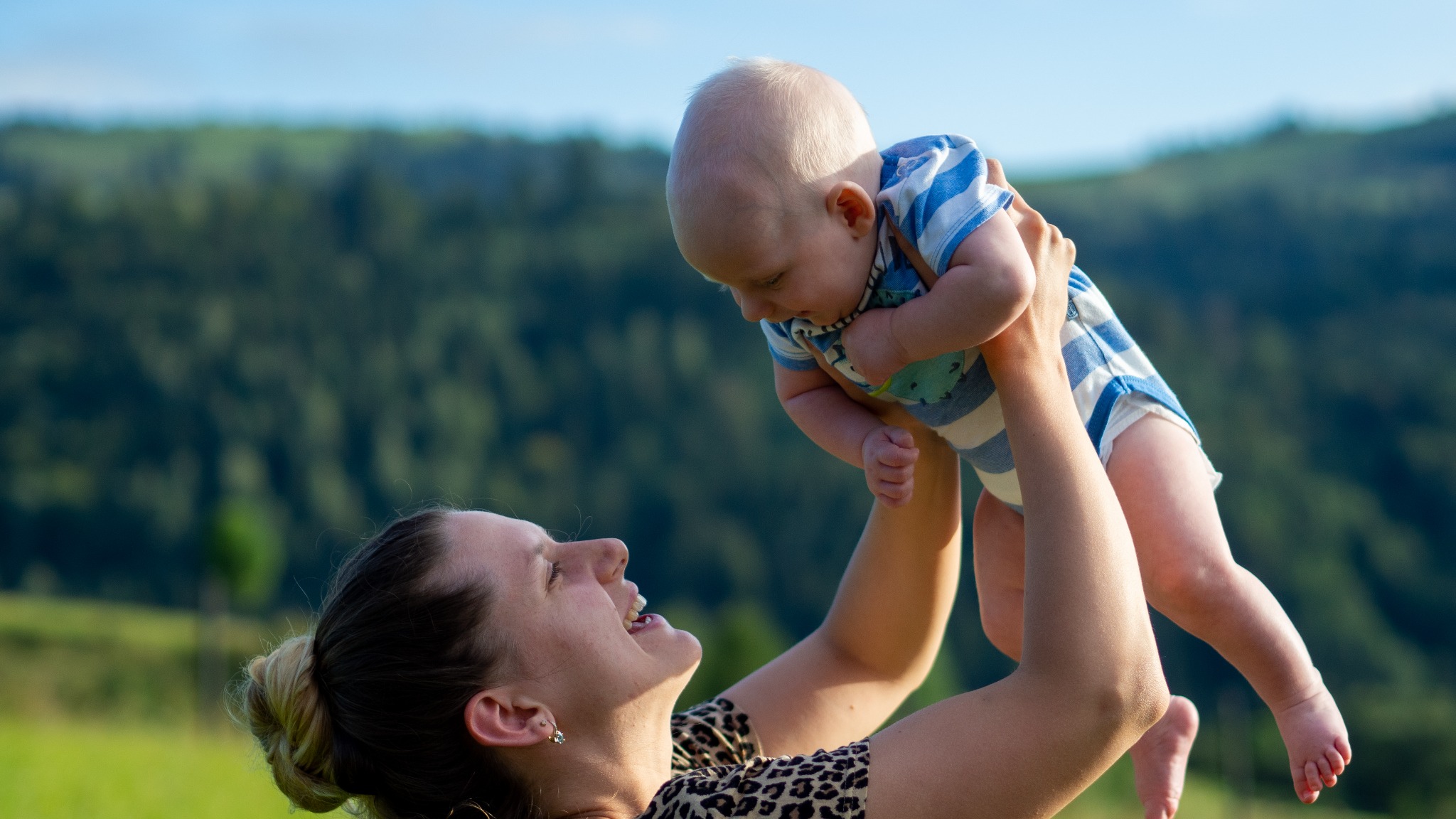 Uśmiechnięta mama podnosi w górę niemowlę w niebiesko-białym pasiastym ubranku na tle zielonych wzgórz