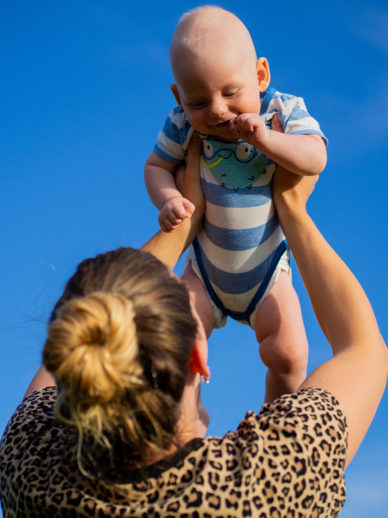 Matka w bluzce w panterkę podrzuca do góry niemowlę w niebiesko-białym body na tle czystego, błękitnego nieba.