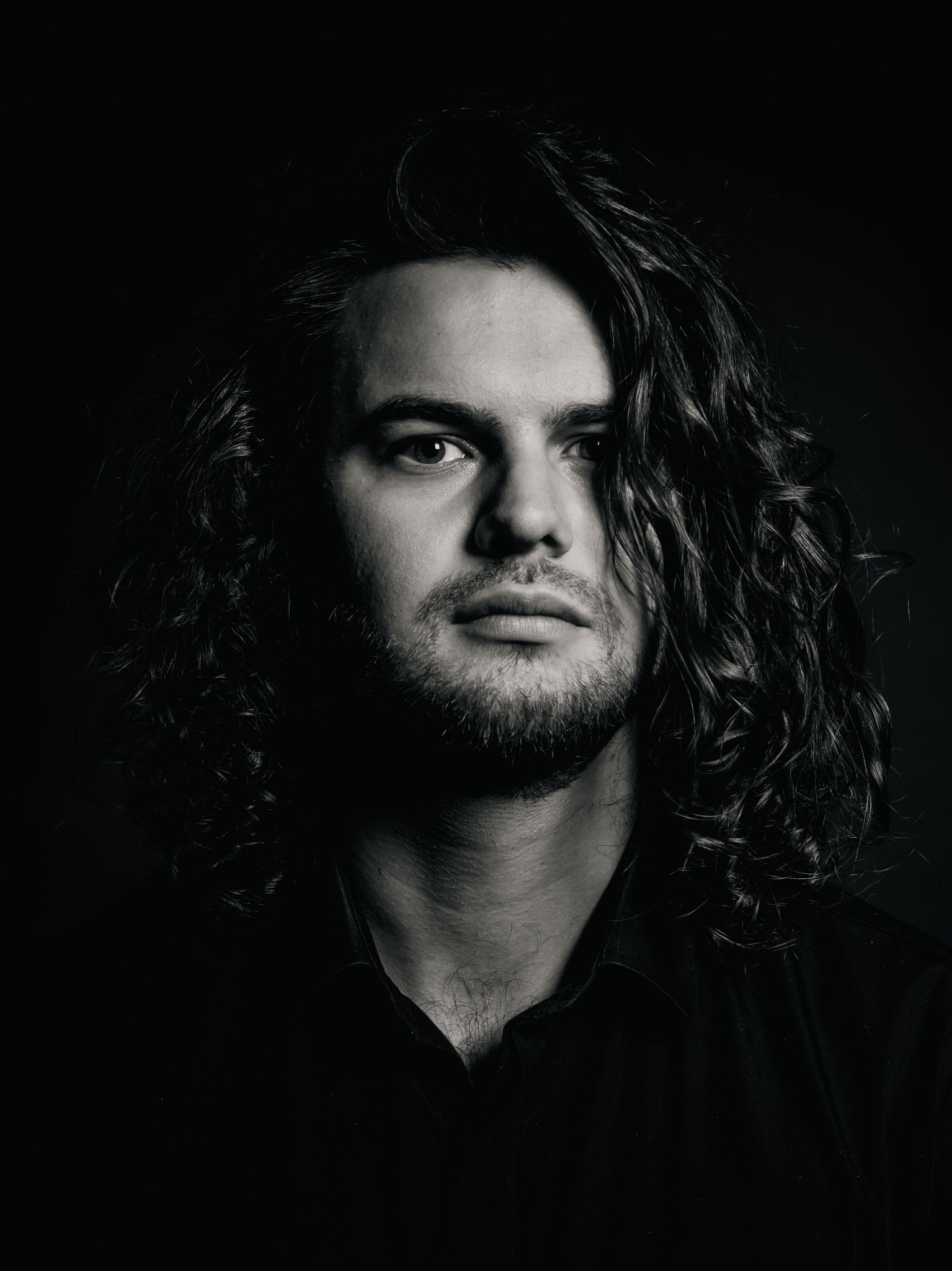 Czarno-biały portret mężczyzny z długimi, kręconymi włosami, zarośniętym zarostem i ciemną koszulą na ciemnym tle.