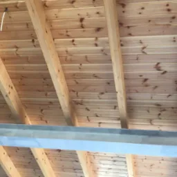 Drewniany sufit z widocznymi belkami i pojedynczym kablem wystającym z desek, wsparty na metalowej belce.