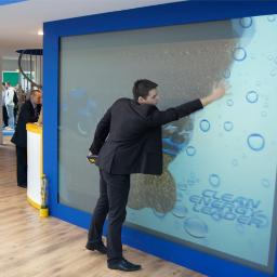 Berrylife-owocna interakcja - Mężczyzna w garniturze wchodzi w interakcję z interaktywną ścianą z motywem czystej energii, widoczne bąbelki i napis 'Clean Energy Leader', w tle fragment ekspozycji targowej z innymi osobami.