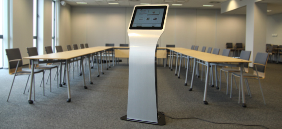 Samostojący infokiosk z ekranem dotykowym w sali konferencyjnej z długimi stołami i krzesłami na kółkach.