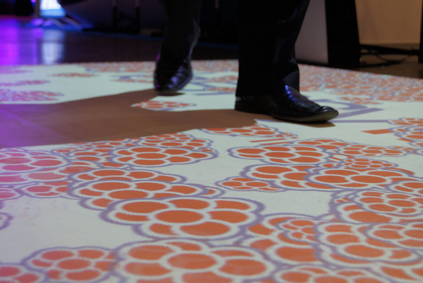 Interaktywna podłoga z projekcją wzoru kwiatowego w odcieniach pomarańczy i fioletu, na której stoi osoba w czarnych butach, rzucająca cień.