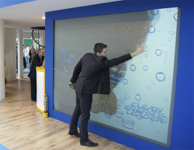 Mężczyzna w garniturze wchodzi w interakcję z interaktywną ścianą z motywem czystej energii, widoczne bąbelki i napis 'Clean Energy Leader', w tle fragment ekspozycji targowej z innymi osobami.