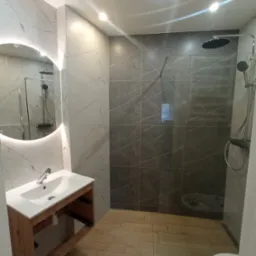 Nowoczesna łazienka z okrągłym lustrem podświetlanym LED, marmurowymi płytkami ściennymi, kabiną prysznicową typu walk-in z ciemnymi płytkami i drewnopodobną podłogą.