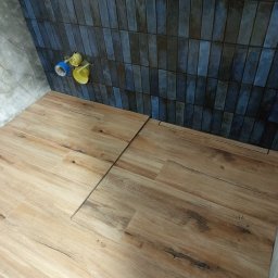 P.W.W.R. PROFESSIONAL Robert Borkowski - Ułożona podłoga z płytek imitujących drewno w łazience z niebieską mozaiką na ścianie i widocznymi elementami instalacji wodnej.
