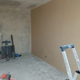 Pokój w trakcie remontu: jedna ściana pomalowana na beżowo, druga surowa, widoczne narzędzia (mieszadło, szpachelki), drabina Krause, sufit z widocznymi łatami gipsowymi.
