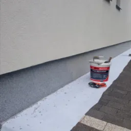 Nakładanie mozaikowego tynku na cokół budynku, wiadro z tynkiem Bau Master i narzędzia obok, zabezpieczona folią podstawa ściany