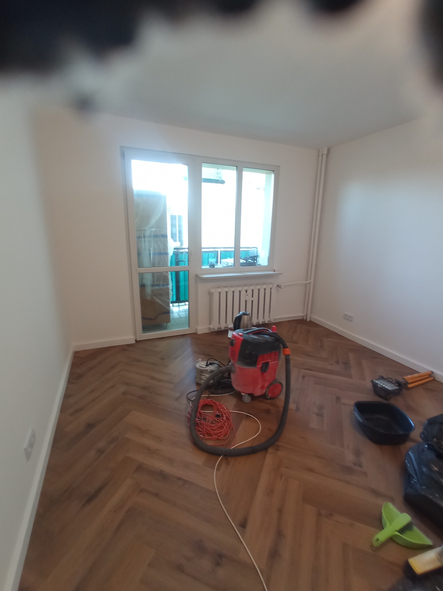 Pokój po ułożeniu paneli podłogowych w jodełkę, widoczny odkurzacz przemysłowy, narzędzia i resztki materiałów budowlanych.