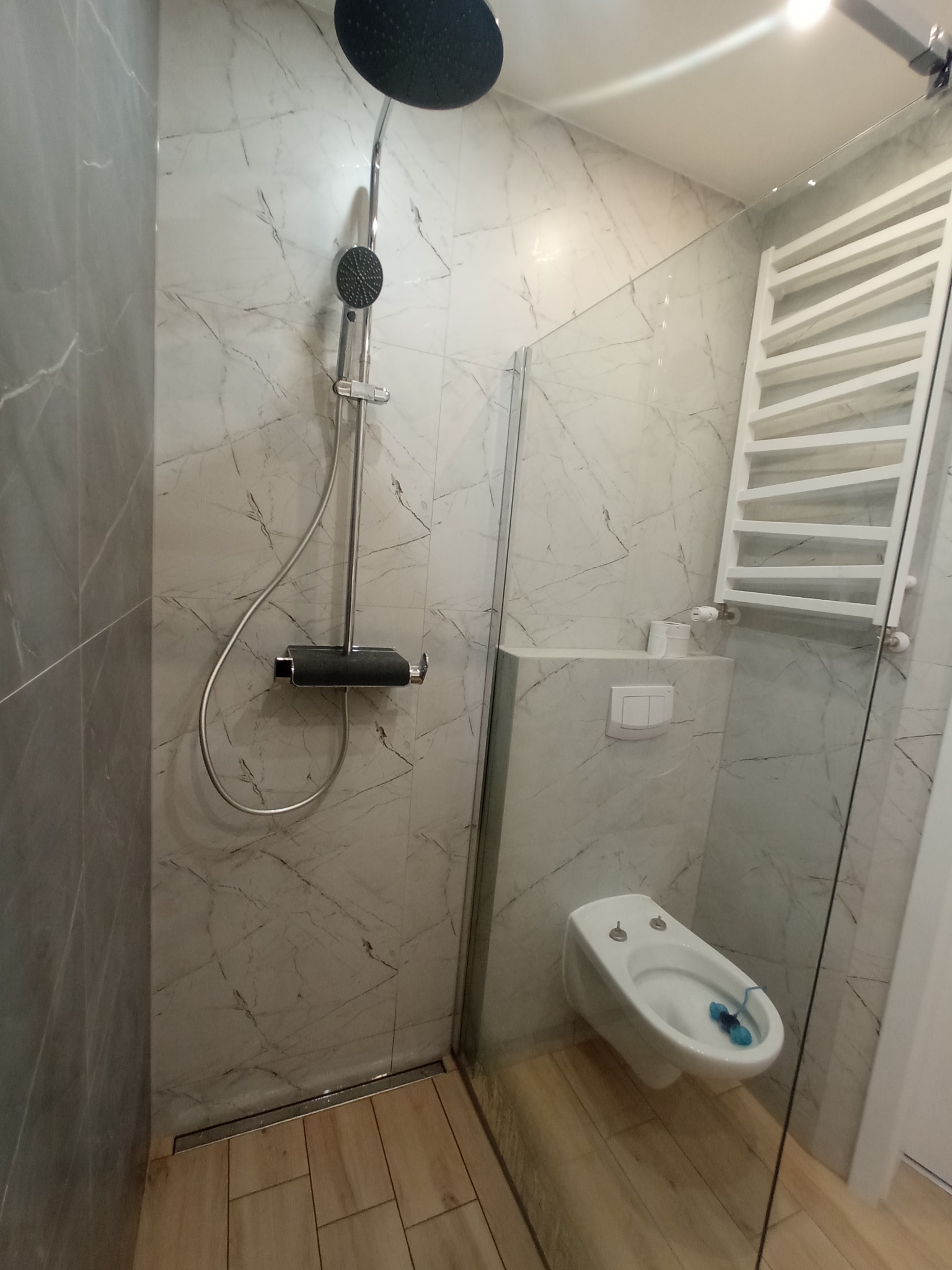 Nowoczesna łazienka z kabiną prysznicową typu walk-in, wykończona płytkami imitującymi marmur, z deszczownicą i bidetem wiszącym. Widoczny grzejnik drabinkowy i drewnopodobna podłoga.