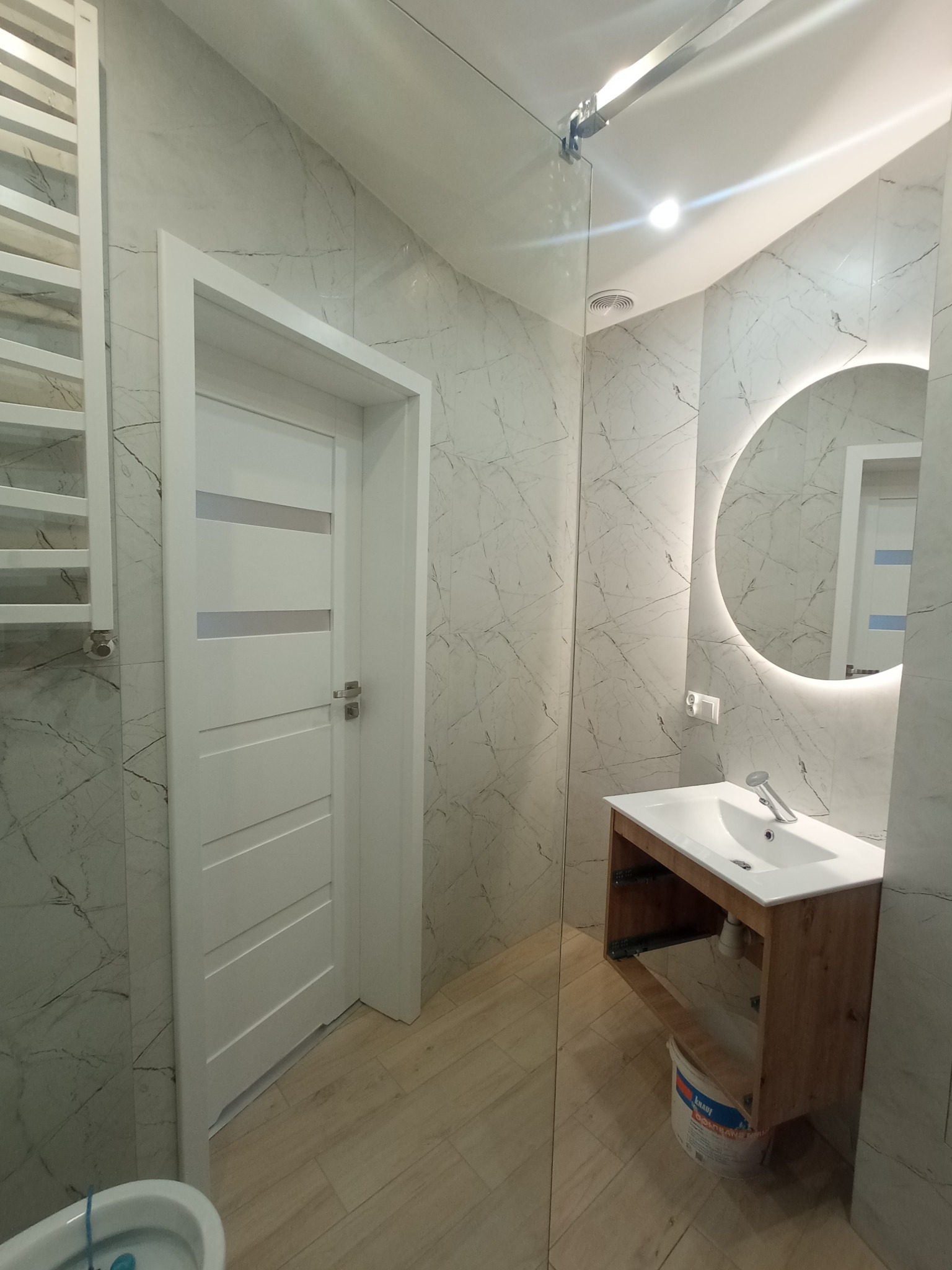 Nowoczesna łazienka z marmurowymi płytkami, oświetlonym okrągłym lustrem, białą umywalką z drewnianą szafką, grzejnikiem drabinkowym i białą ścianą działową ze szklanym panelem. Na podłodze panele...