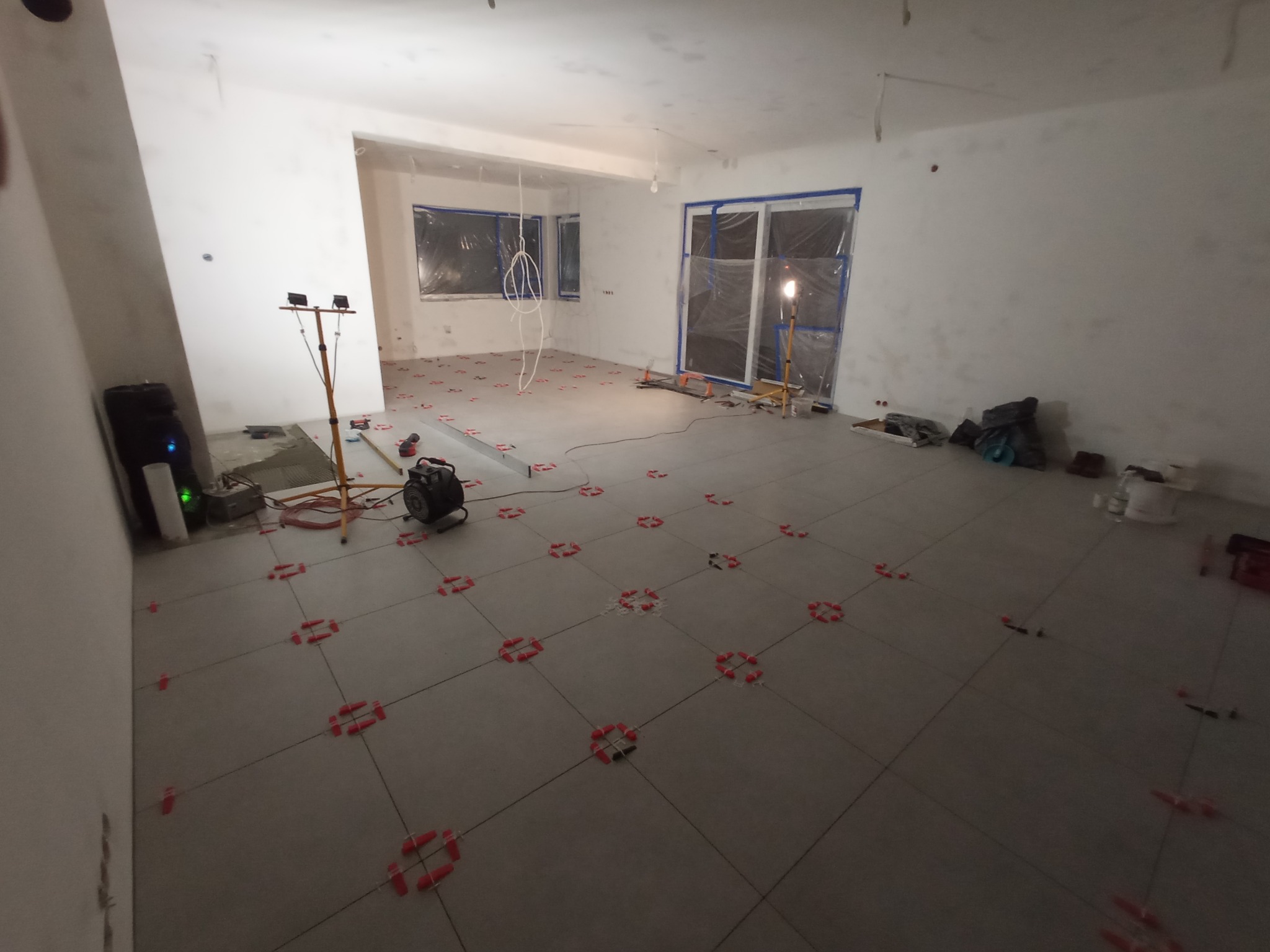 Szare płytki podłogowe ułożone w dużym pokoju, z czerwonymi krzyżykami dystansowymi, okna i drzwi balkonowe zabezpieczone folią malarską, widoczne narzędzia i lampa budowlana.