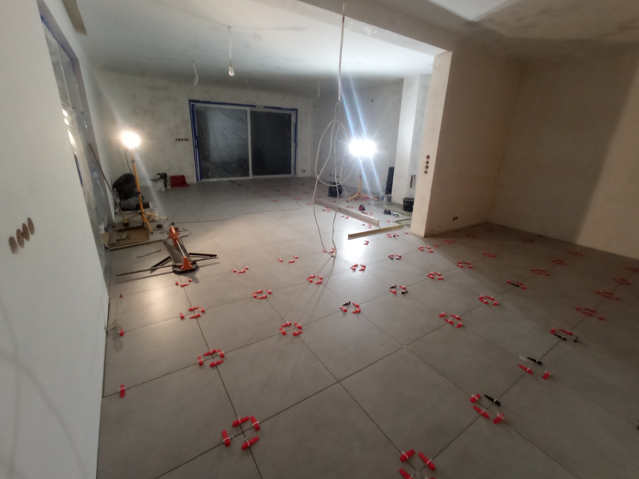Szare płytki podłogowe w trakcie układania, z czerwonymi i czarnymi krzyżykami dystansowymi, widoczne narzędzia i oświetlenie budowlane, przygotowane do fugowania.