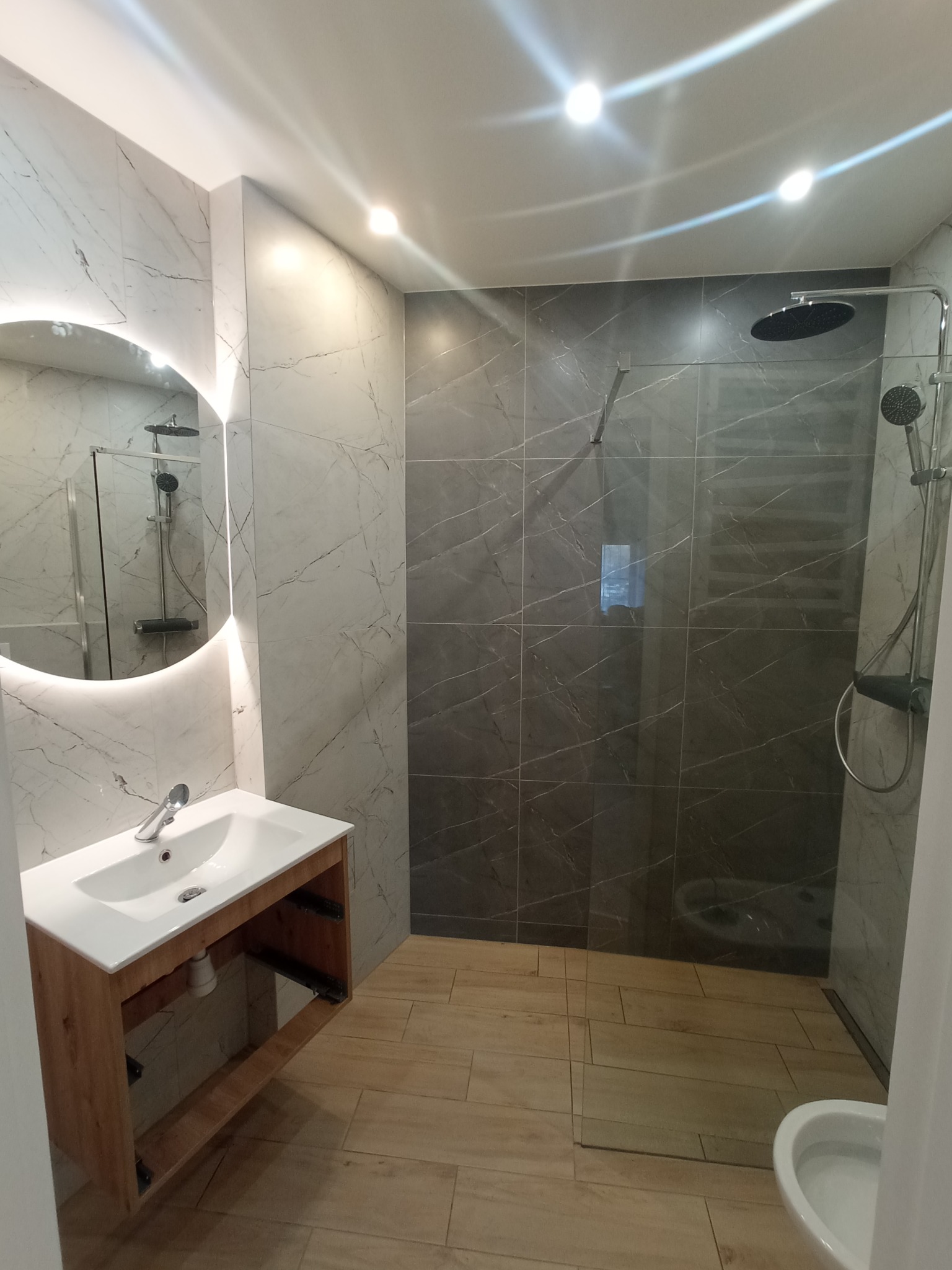 Nowoczesna łazienka z okrągłym lustrem podświetlanym LED, marmurowymi płytkami ściennymi, kabiną prysznicową typu walk-in z ciemnymi płytkami i drewnopodobną podłogą.