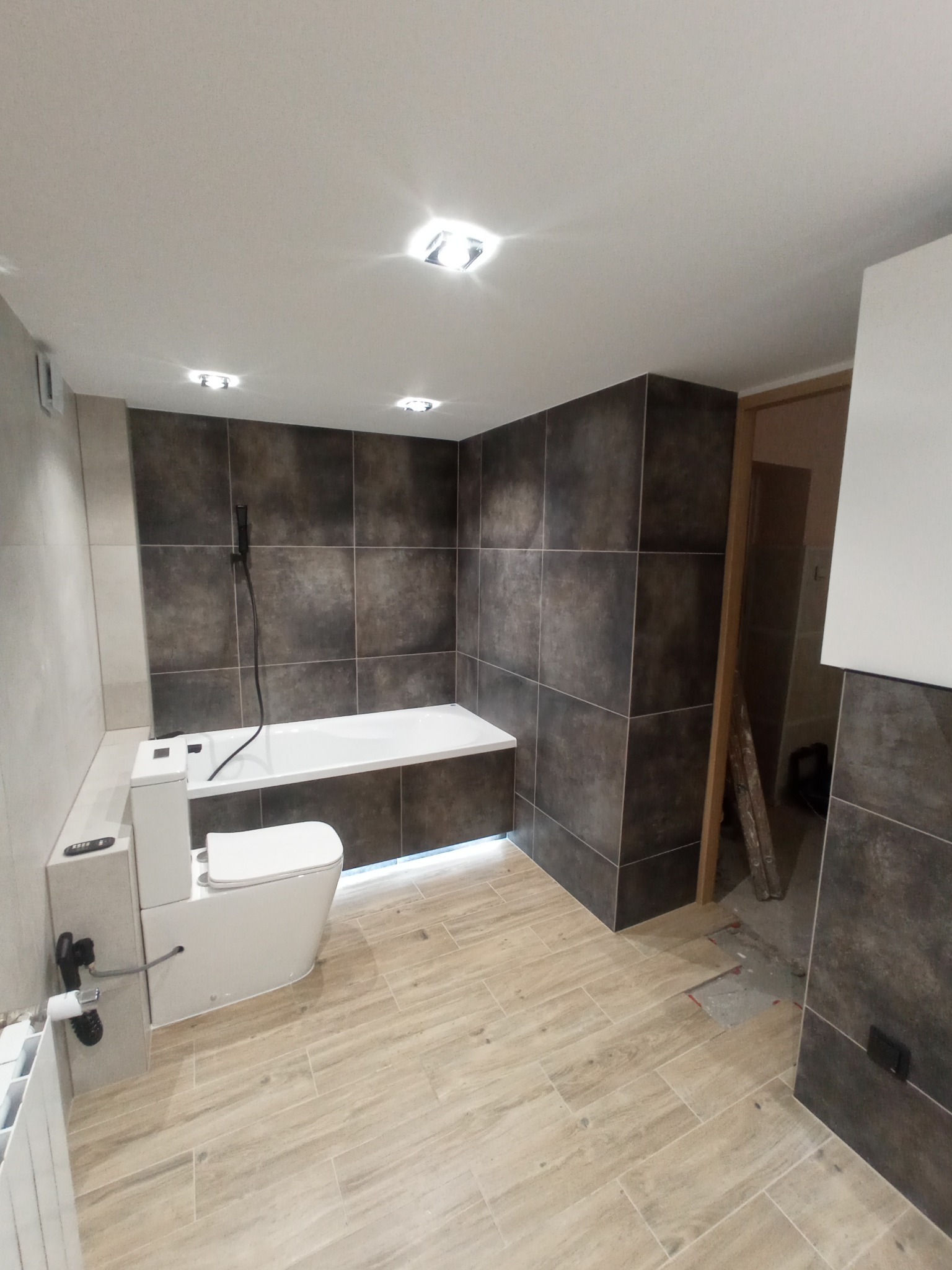 Wykończona łazienka z brązowymi i beżowymi płytkami, podwieszana toaleta z panelem sterującym, biała wanna z czarną słuchawką prysznicową, drewnopodobna podłoga, sufit z oświetleniem punktowym.