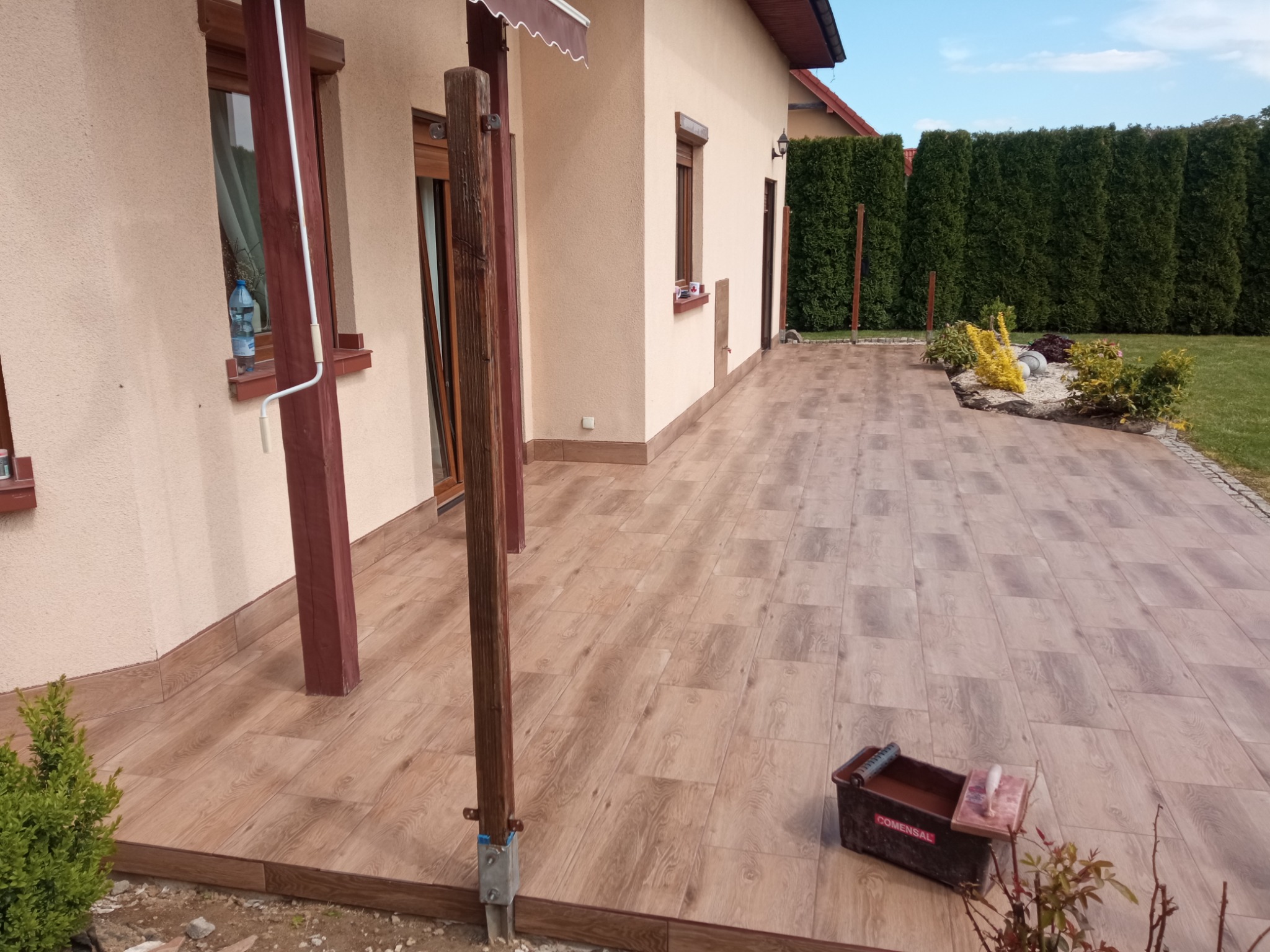 Ułożona, drewnopodobna terakota na tarasie przy domu. Widoczny pojemnik z zaprawą i narzędzia glazurnicze. Tło: krzewy i błękitne niebo.