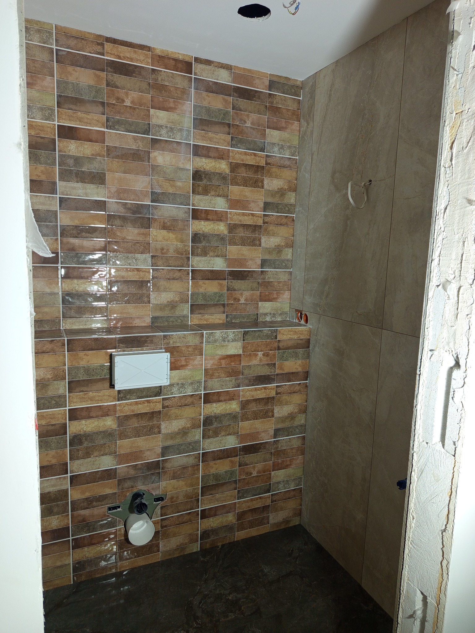 Wykończona ściana w łazience z brązowymi, prostokątnymi płytkami, obok ściana z beżowych płytek imitujących marmur. Widoczne elementy instalacji sanitarnej.