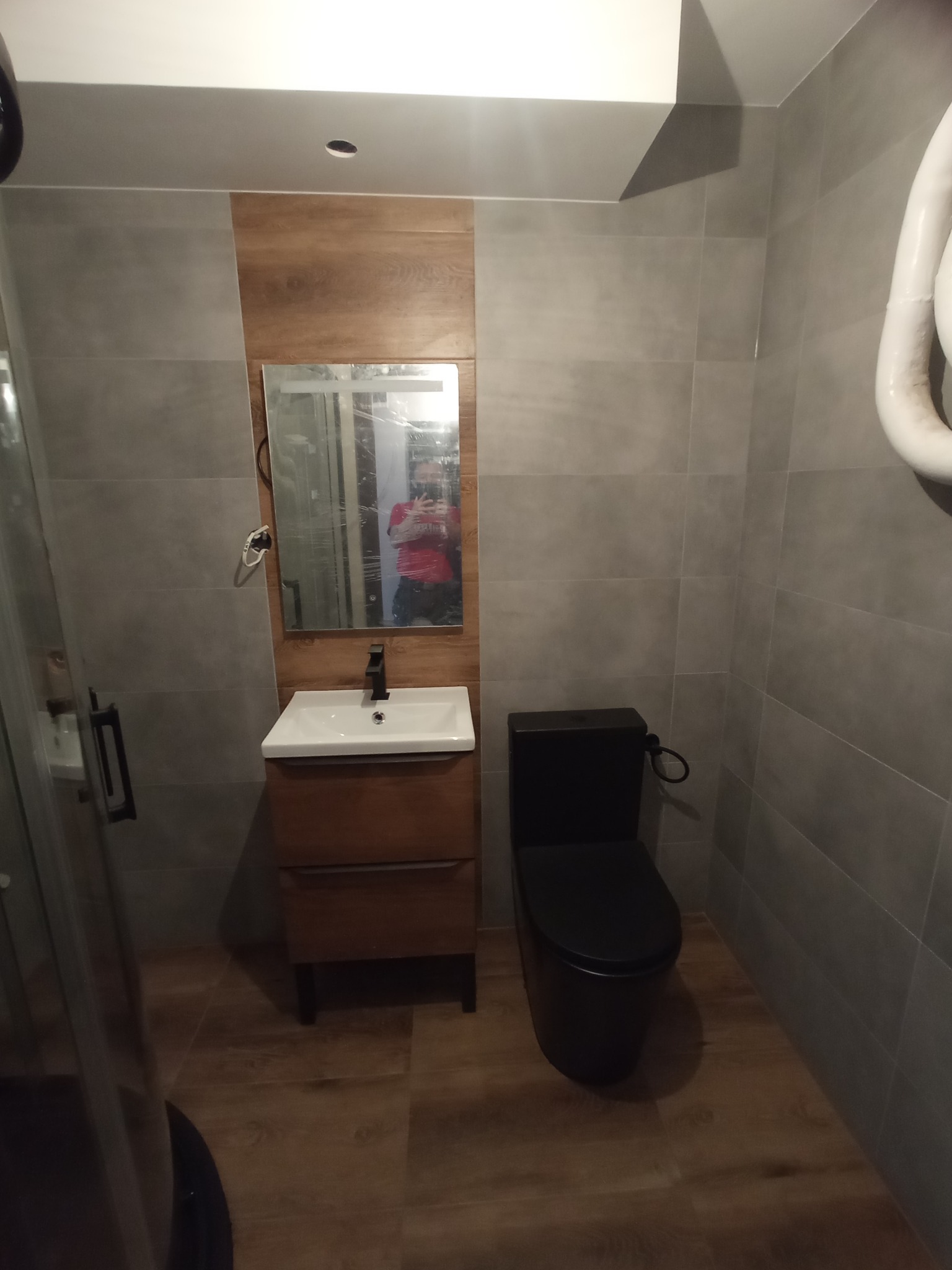 Wykończona łazienka z szarymi płytkami, drewnopodobną ścianą, umywalką z szafką i czarną toaletą. Widoczne odbicie osoby w lustrze.