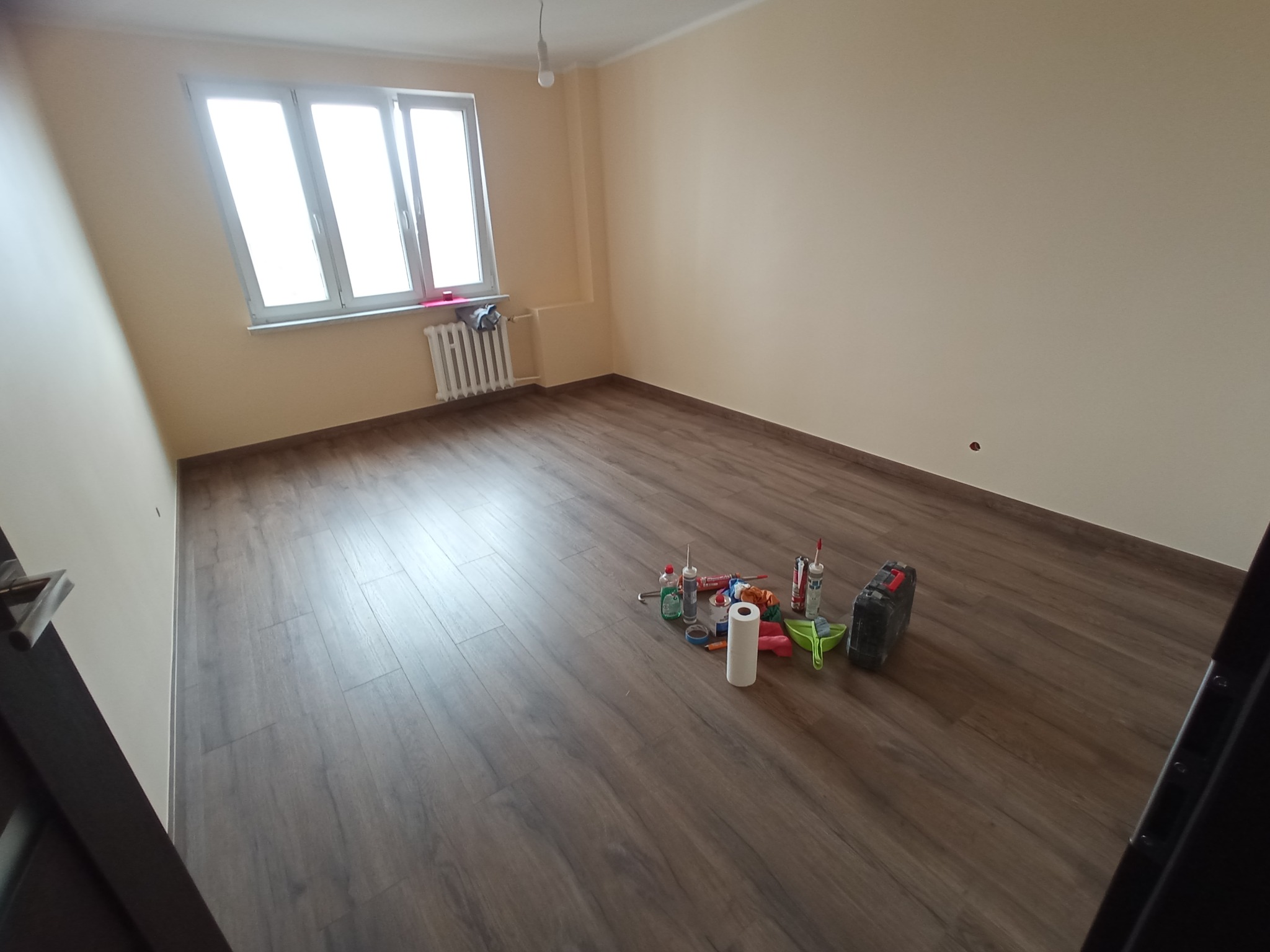 Jasne wnętrze po remoncie: brązowe panele podłogowe, beżowe ściany, okno z grzejnikiem. Na podłodze narzędzia i materiały budowlane.