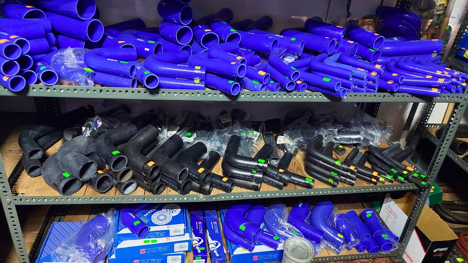 Półki regału warsztatowego wypełnione niebieskimi i czarnymi gumowymi wężami samochodowymi, w tle widoczne opakowania producenta.