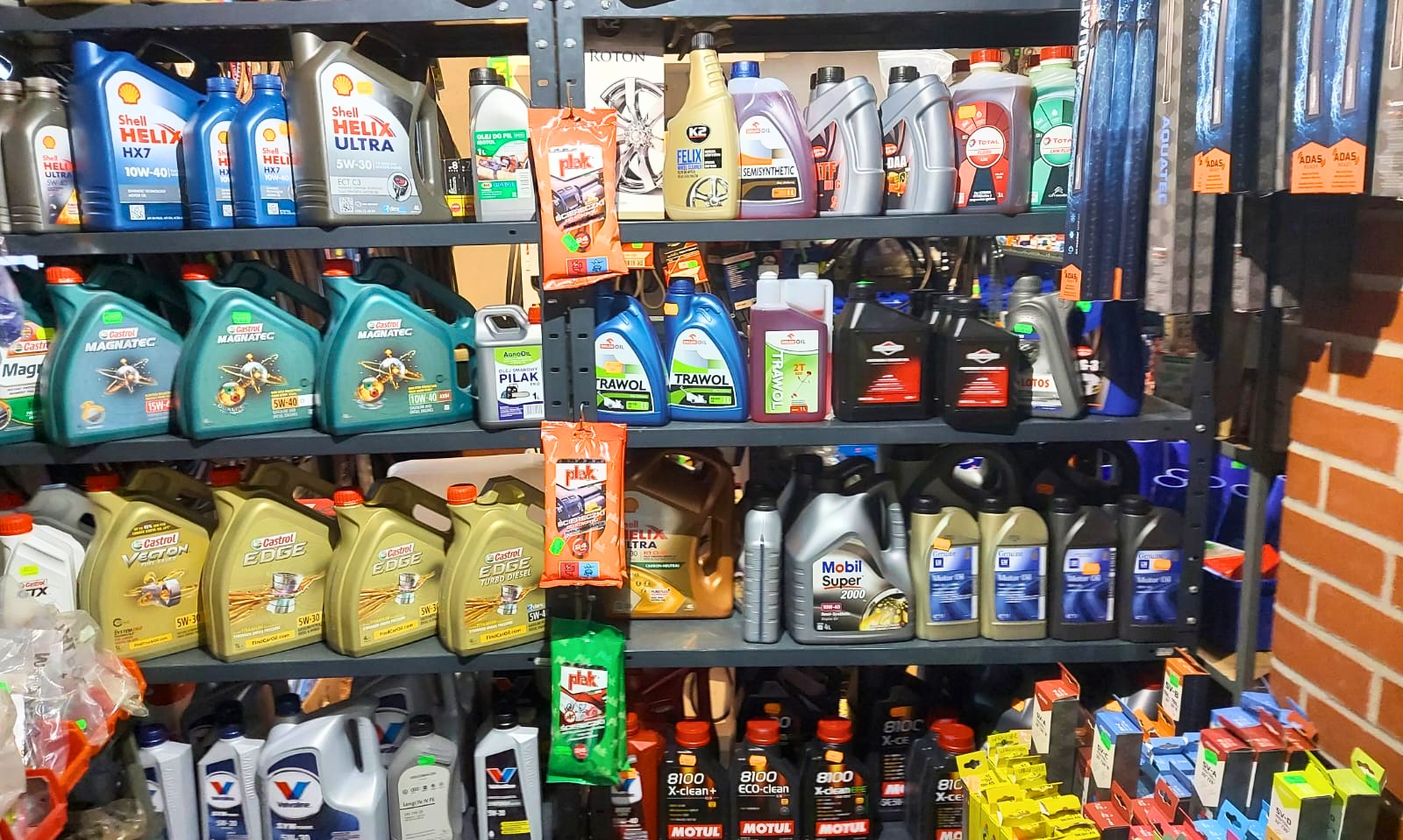 Półki warsztatu samochodowego wypełnione różnorodnymi olejami silnikowymi różnych marek i specyfikacji, w tym Shell Helix, Castrol Magnatec i Edge, Mobil Super, Motul 8100, oraz preparatami...