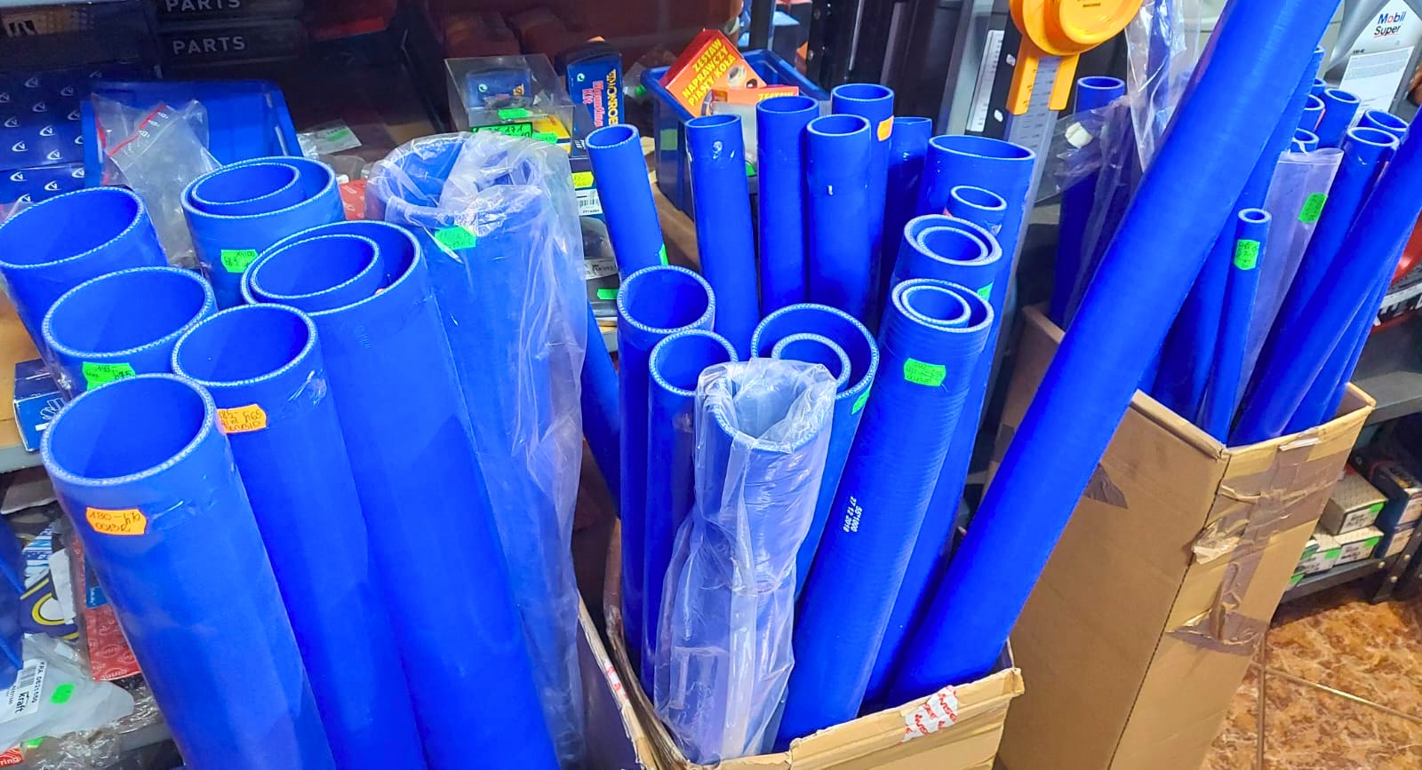 Magazyn niebieskich, silikonowych węży samochodowych o różnych średnicach, przechowywanych w kartonowych pudłach, z widocznymi etykietami cenowymi i opakowaniami części zamiennych w tle.