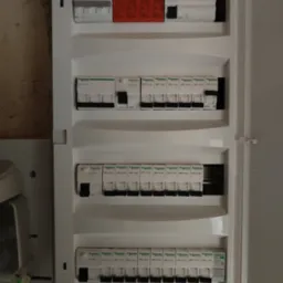 Otwarta skrzynka elektryczna z zamontowanymi bezpiecznikami i wyłącznikami różnicowoprądowymi marki Schneider, widoczne oznaczenia amperażu i modelu, na ścianie obok widoczny fragment instalacji.