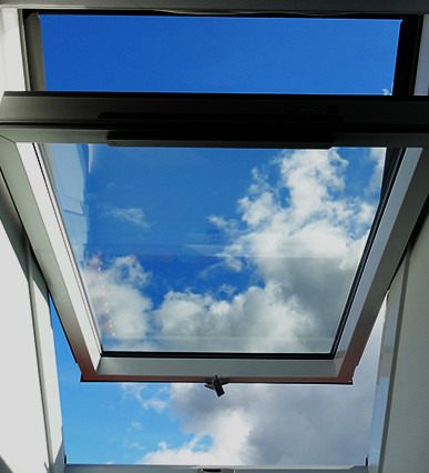 Otwarty okno dachowe z widokiem na błękitne niebo i chmury, uchwyt okna w pozycji otwartej.