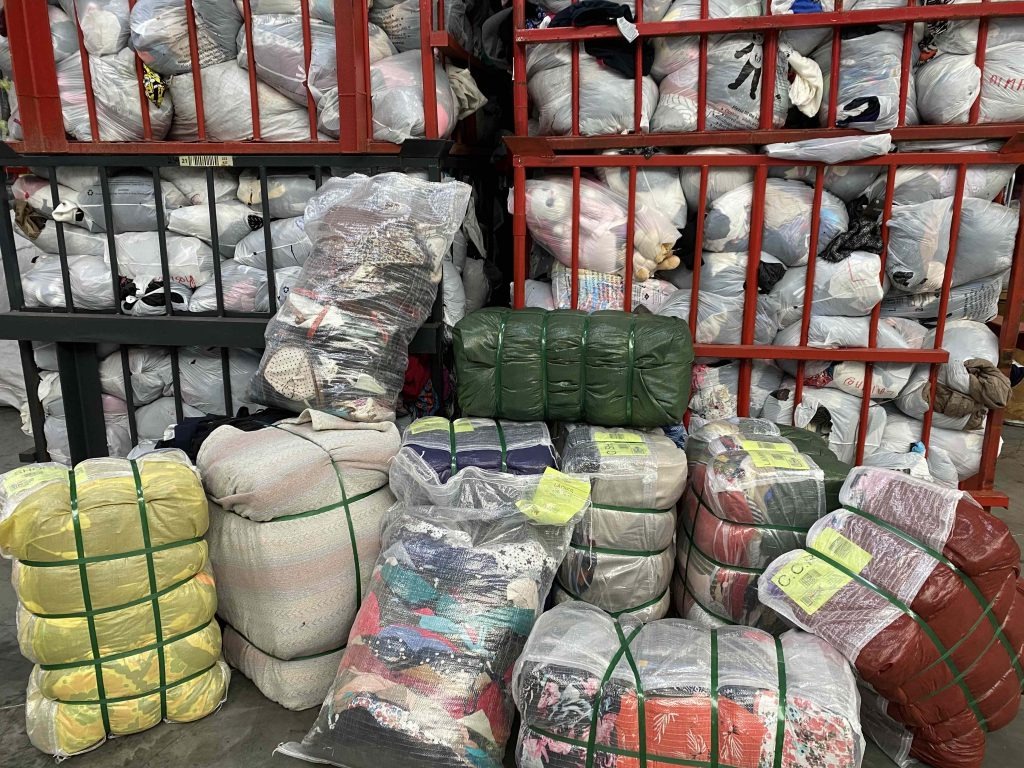 Stosy sprasowanej, używanej odzieży w workach i paczkach, składowane na metalowych regałach w magazynie.