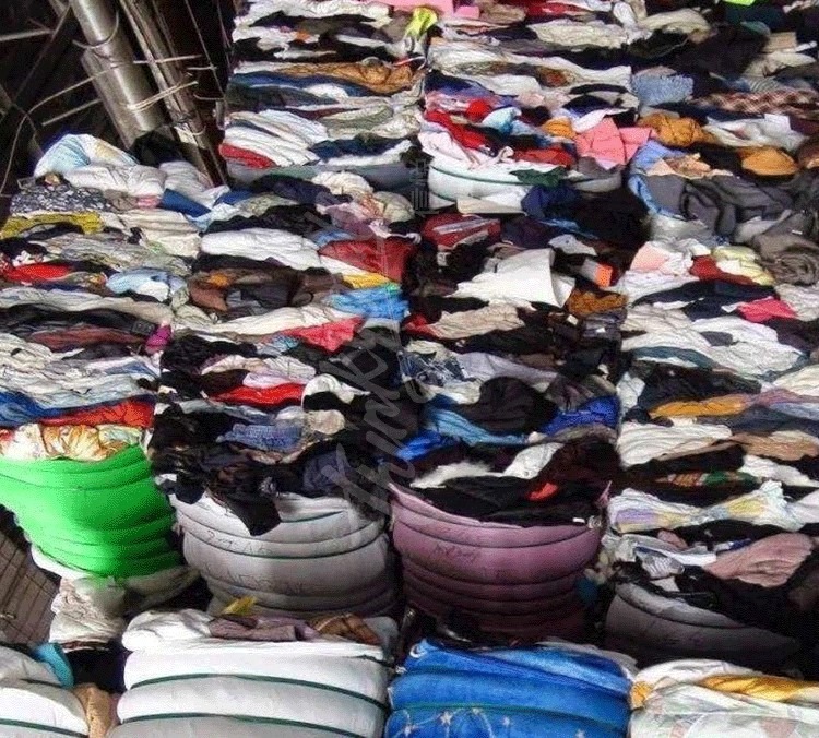 Stosy sprasowanej odzieży w belach, widoczne różne kolory i faktury materiałów, oświetlenie magazynowe.