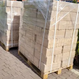 Dwa palety brykietu drzewnego ułożone na kostce brukowej, owinięte folią i zabezpieczone taśmami.