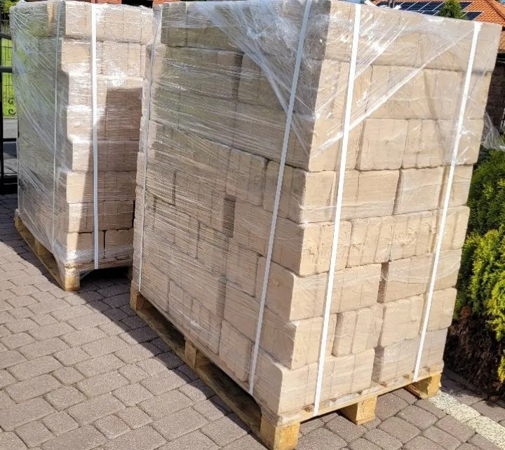 Dwa palety brykietu drzewnego ułożone na kostce brukowej, owinięte folią i zabezpieczone taśmami.