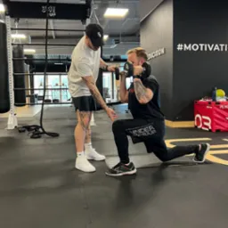 Mężczyzna w pozycji wykroku z hantlą trzymaną przed sobą, nadzorowany przez drugiego mężczyznę w siłowni; widoczny napis '#MOTIVATION' na ścianie.