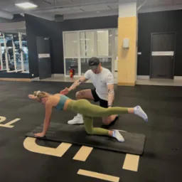 Kobieta wykonuje ćwiczenie na macie w siłowni, mężczyzna z tatuażami na rękach koryguje jej pozycję.