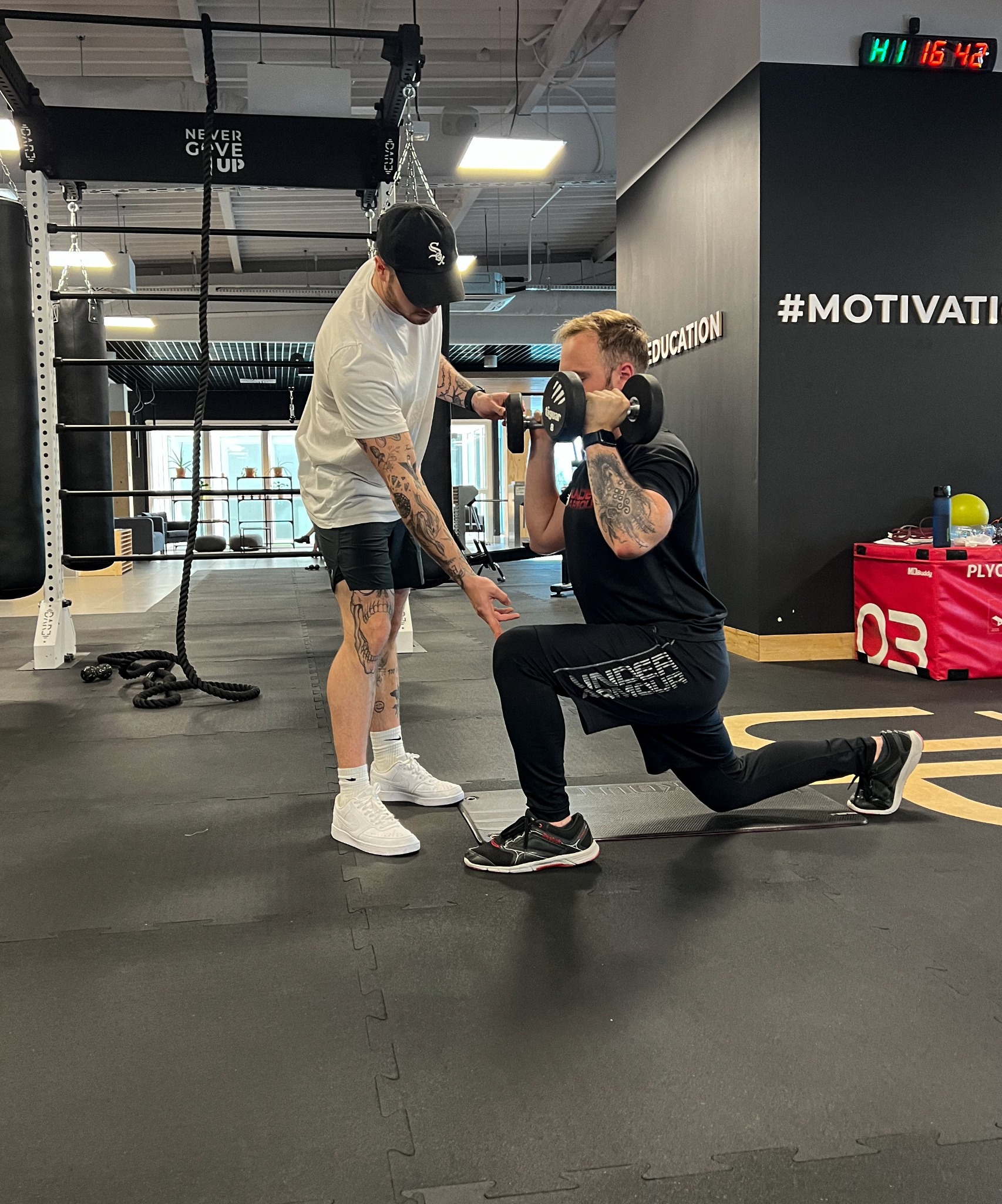 Mężczyzna w pozycji wykroku z hantlą trzymaną przed sobą, nadzorowany przez drugiego mężczyznę w siłowni; widoczny napis '#MOTIVATION' na ścianie.