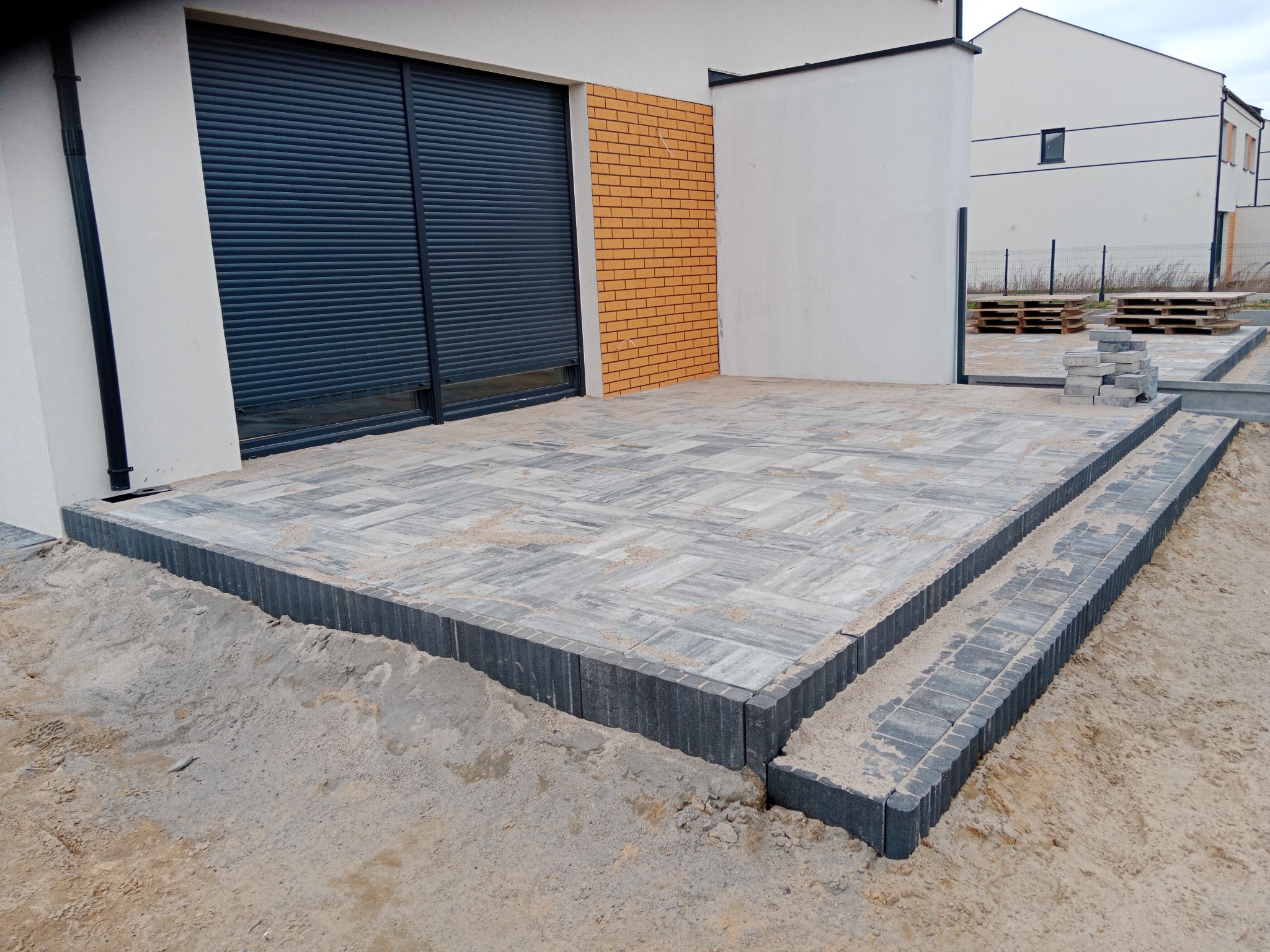 Nowo ułożony taras z szarej kostki brukowej z dwoma stopniami, widoczne obrzeża i piasek, w tle budynek z roletami antywłamaniowymi i palety z kostką.