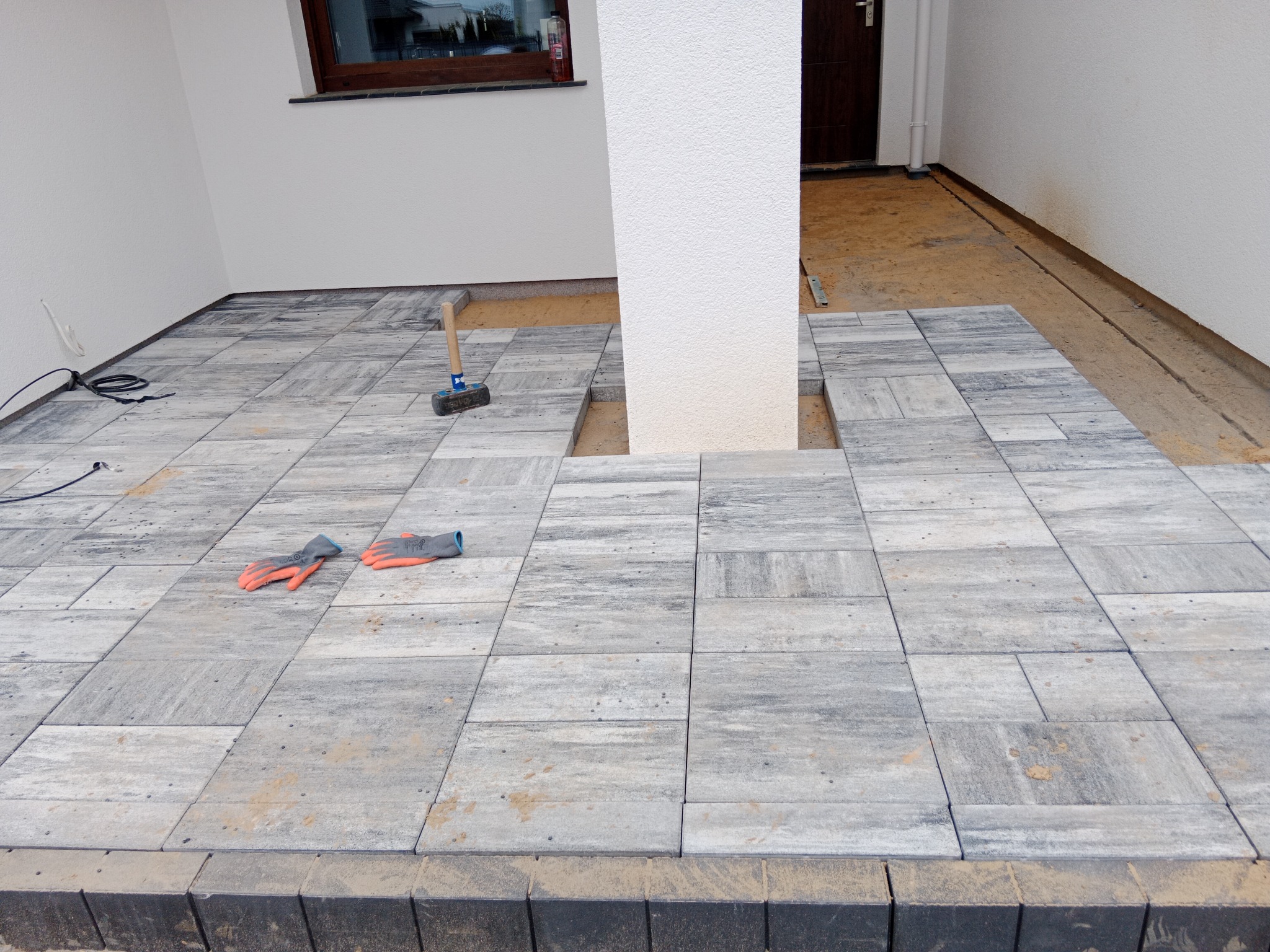 Fragment tarasu z szarych kostek brukowych w trakcie prac, widoczny gumowy młotek i robocze rękawice na powierzchni, słup podtrzymujący konstrukcję domu z wyciętym miejscem na kostkę, tło stanowi...