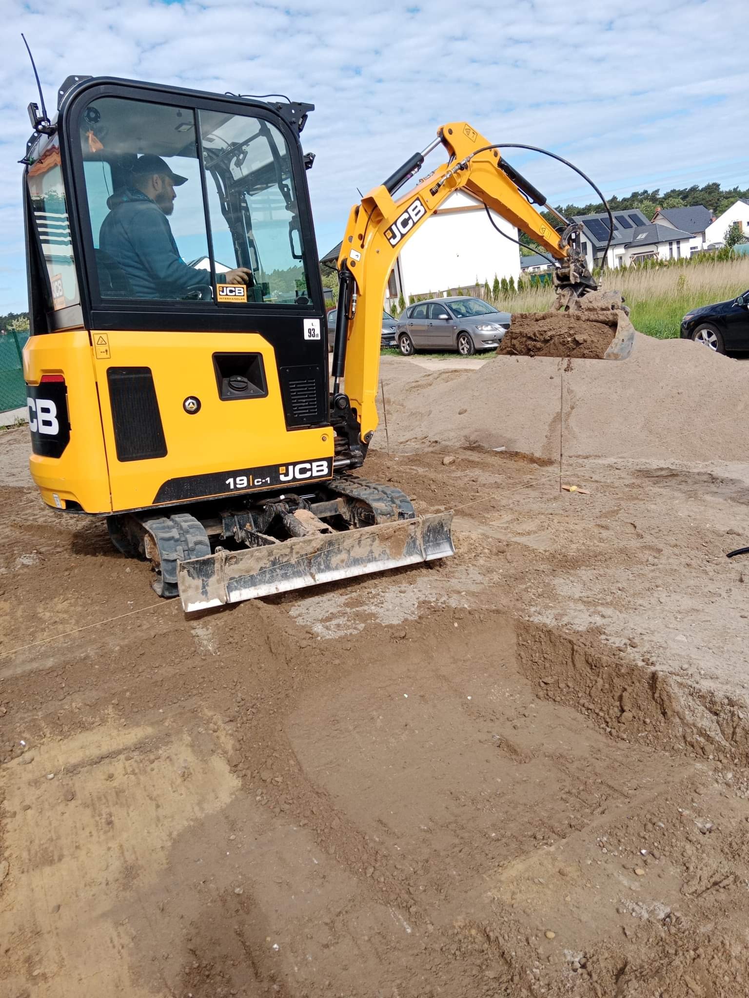 Żółta minikoparka JCB 19C-1 z operatorem w kabinie nabiera ziemię, widoczne wykopane fundamenty, w tle domy jednorodzinne