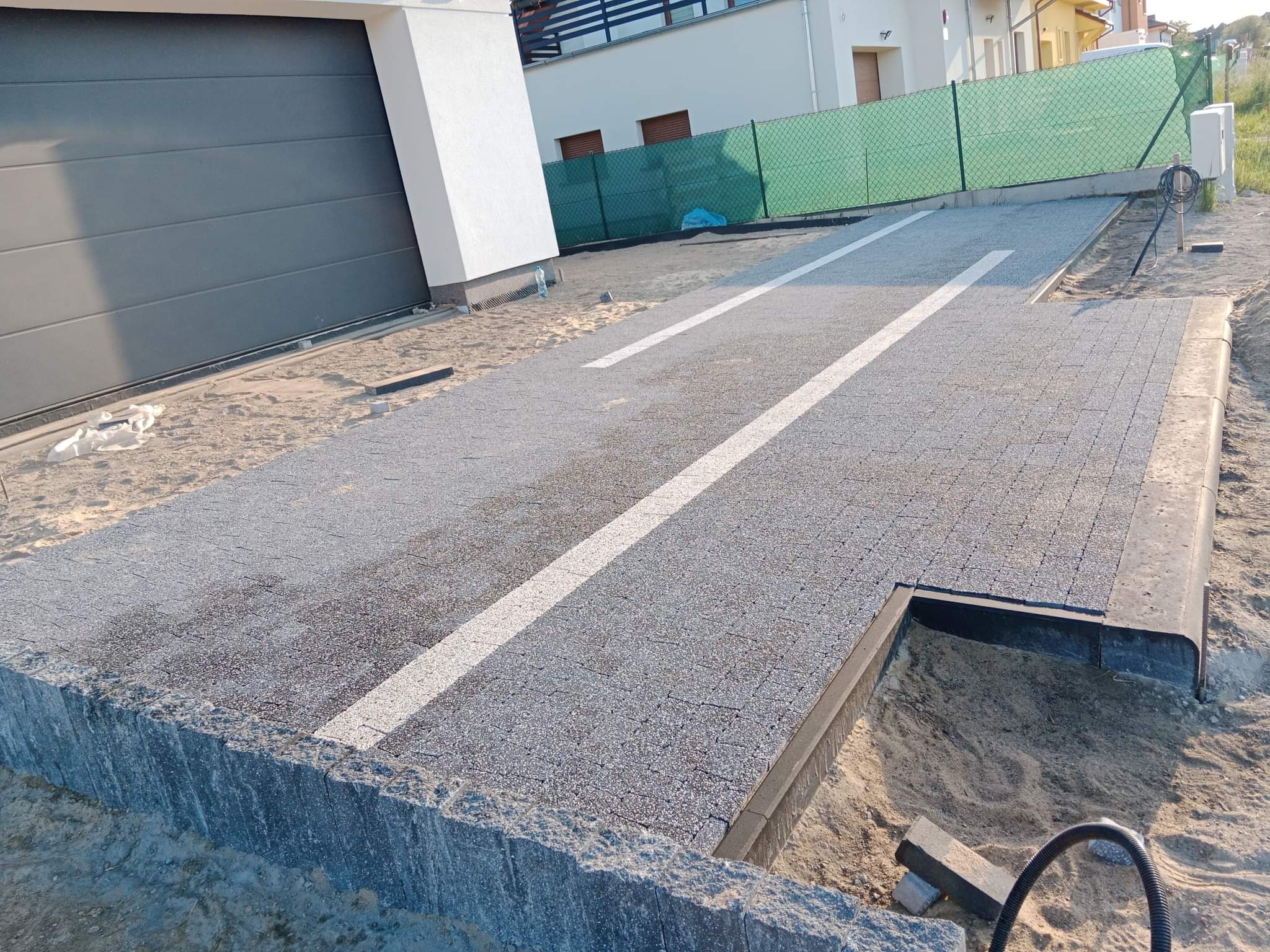 Nowo ułożona szara kostka brukowa z białymi pasami, tworząca podjazd przed garażem w nowoczesnym domu jednorodzinnym, widoczne krawężniki i piasek po bokach.